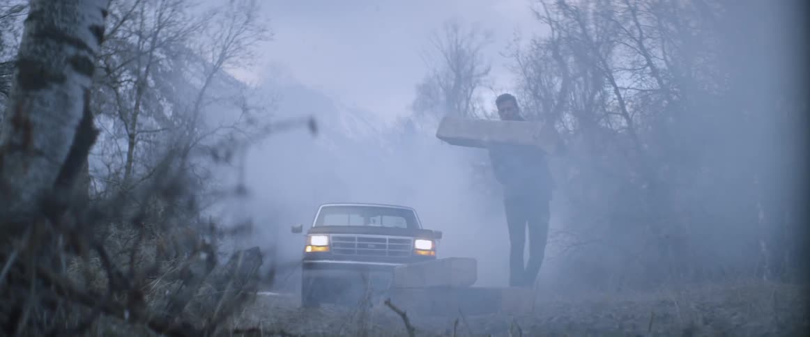 A man is dropping a wood beam outside at dusk while it snows