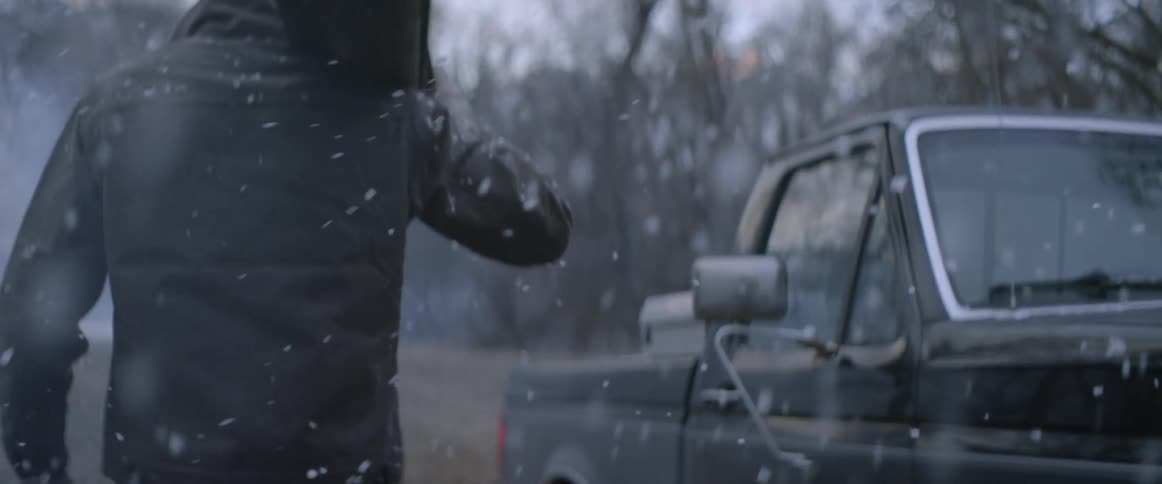 Snow falls on a man carrying a piece of wood