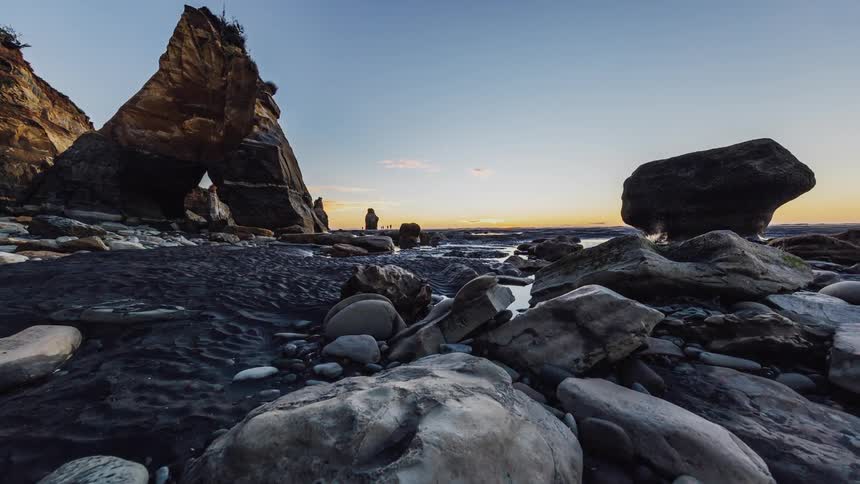 People are walking on a rocky beach as the sun is setting