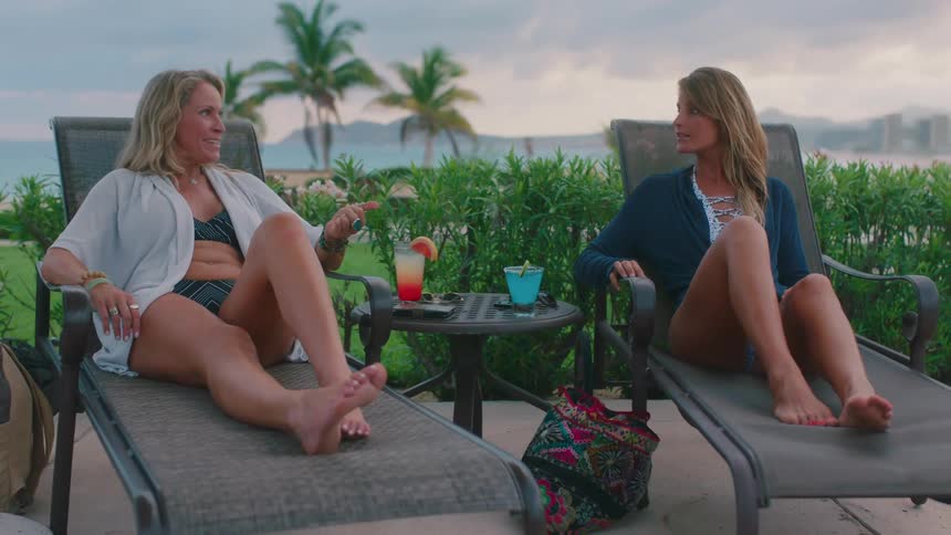 Two women sit in poolside lounge chairs and talk