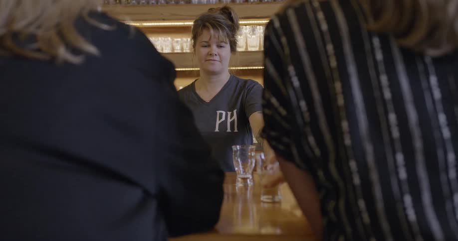 A person puts a glass down on the bar and the bartender turns to get another drink