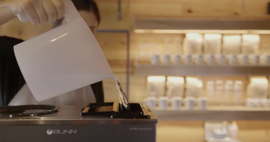 A woman is pouring water into a coffee maker in a coffee shop
