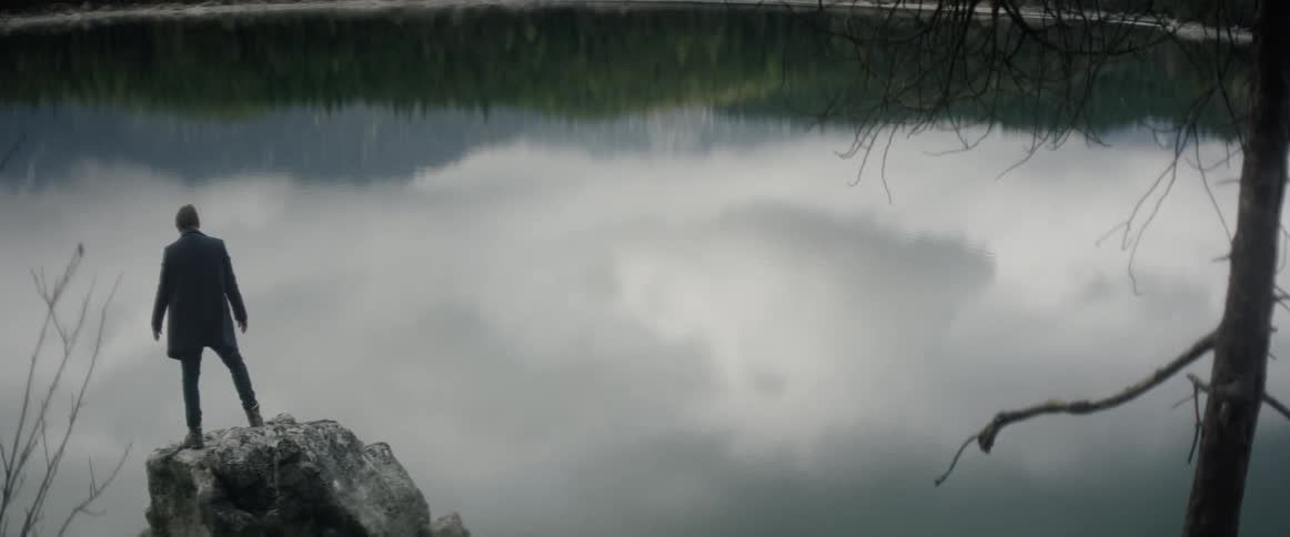 Man climbing up and standing on rock overlooking lake with reflection of clouds