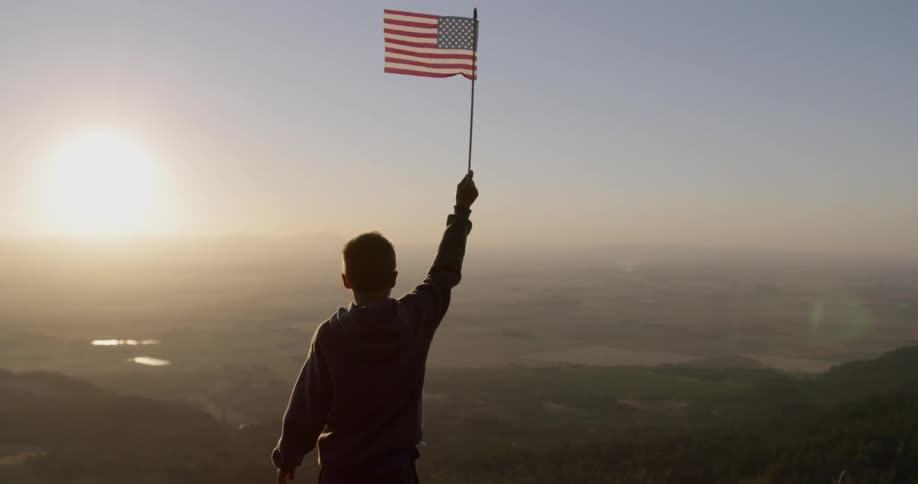 A boy holds an american flag in mountains at sunset