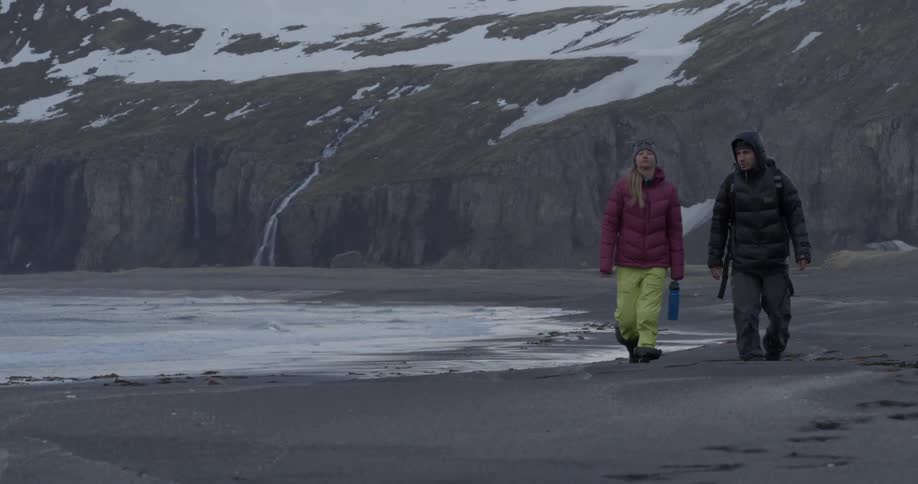 Two people are walking along the beach with a snow covered hill behind them