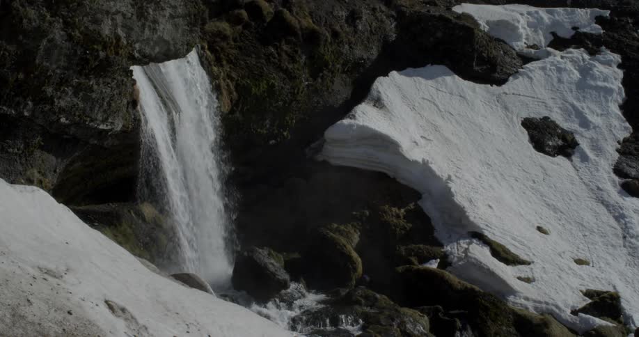 A waterfall is cascading over a ledge and down a snow-covered hillside during the day