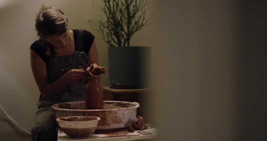 A woman is forming a piece of clay on a pottery wheel inside during the day