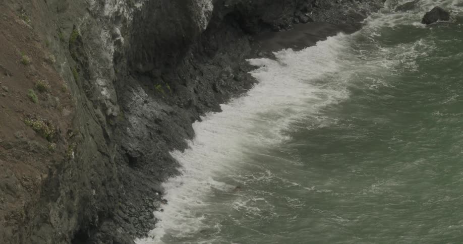 An overhead view of the ocean hitting the coast and bottom of a large cliff