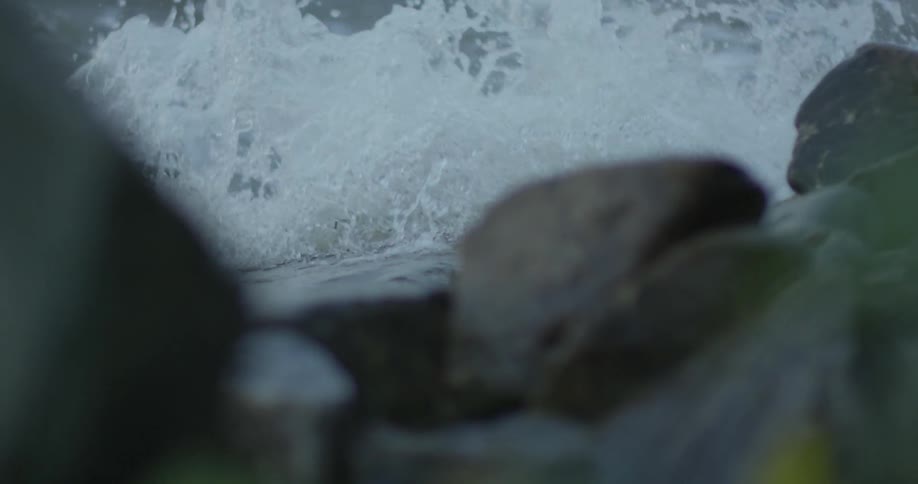 A wave is cresting and breaking over rocks on the shore during the day