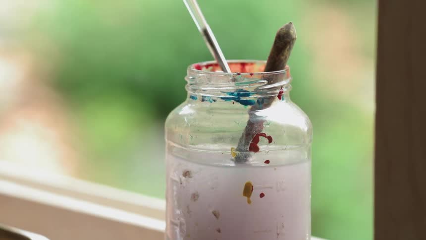 A painter swirls a brush in a jar of water to clean it off