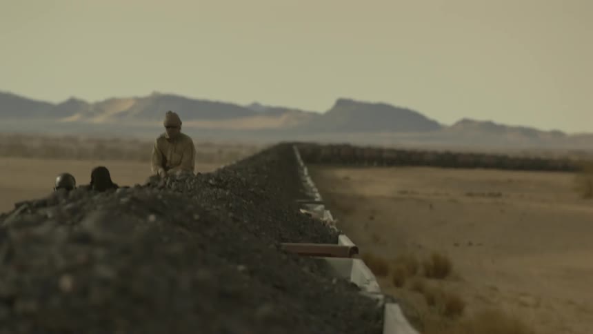 People are riding atop open gravel cars on a train going through the desert