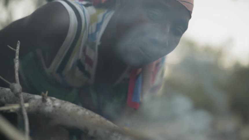 An African woman is blowing on a fire to help it start outside at dusk