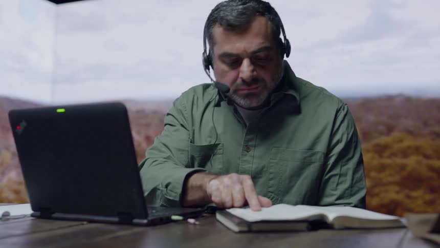 A man works at a desk with imagery being projected on the wall behind him