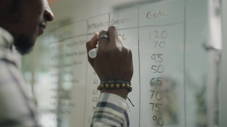 Man erasing a number on a glass board and smiling after being handed a piece of paper from another man