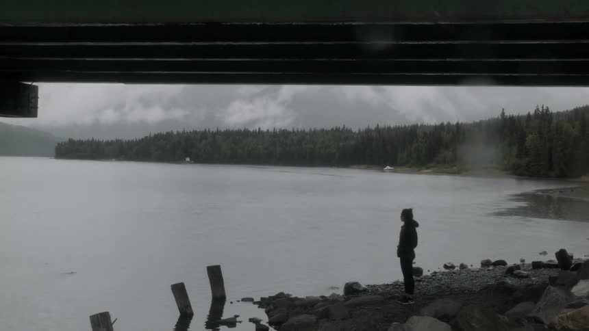 A woman is standing on a shoreline and looking around on a cold day