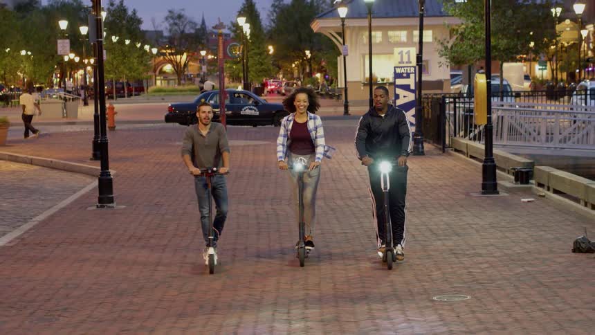 Three friends are riding scooters in a city at dusk