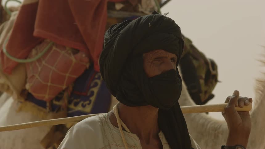 A man wearing a headscarf stands in front of a saddled camel