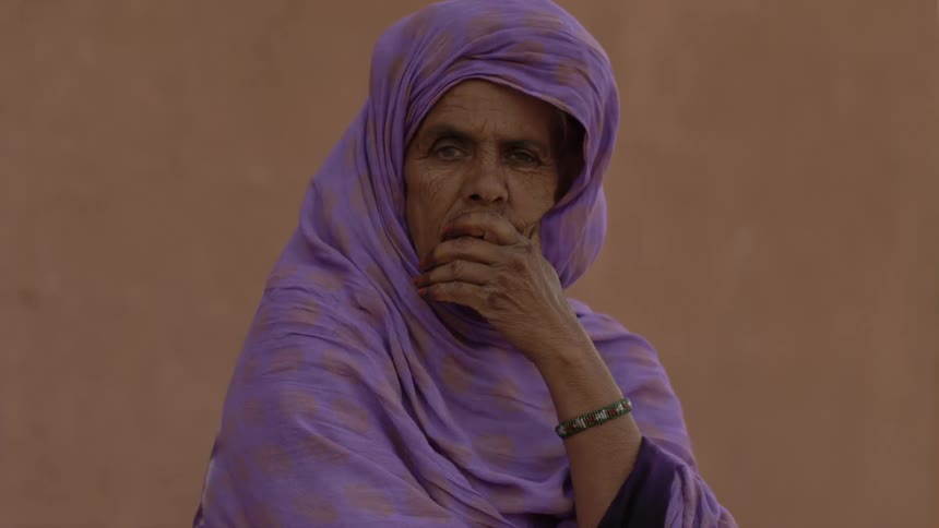 A woman wearing a headscarf sits outside with her hand over her mouth