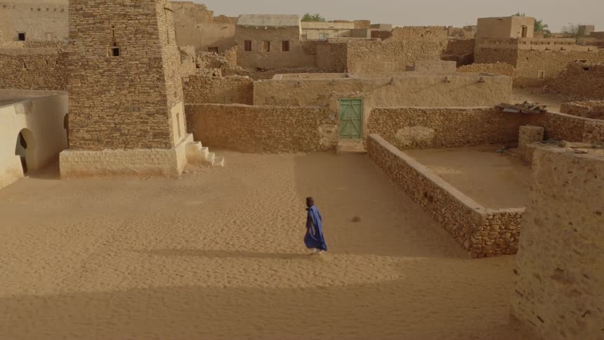 A man wearing blue robes is walking in an old village in the desert