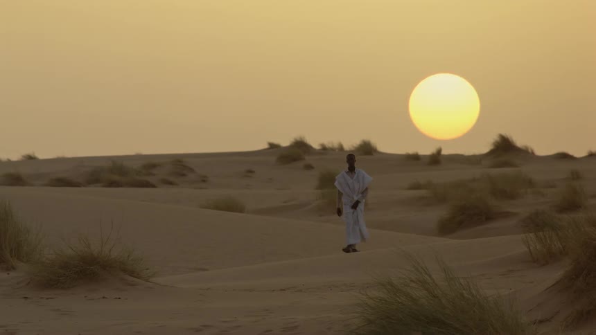 A man is walking over sand dunes in a desert at sunset