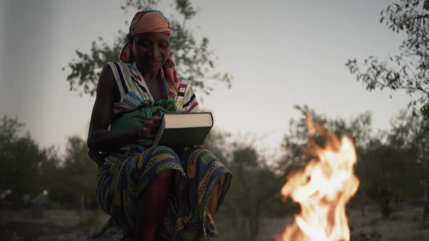 An African woman opening a book as she sits by a fire