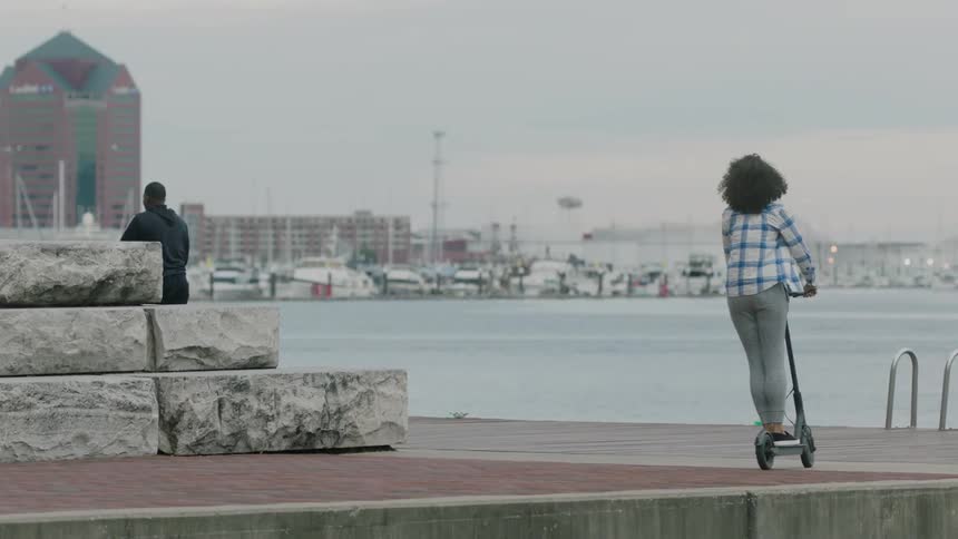 People riding kick scooters in an area next to some water in a city