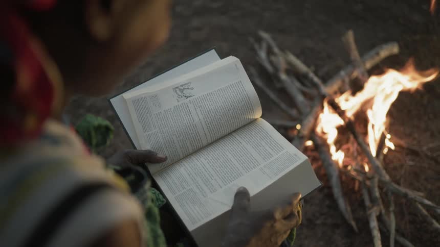 A woman sitting outside at dusk near a campfire is reading a bible
