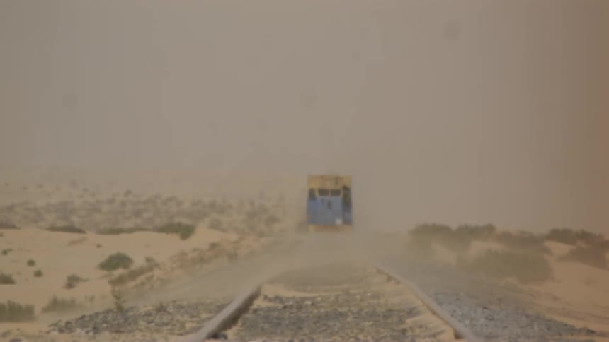 Dust is blowing over train tracks in a desert landscape as a train approaches during the day