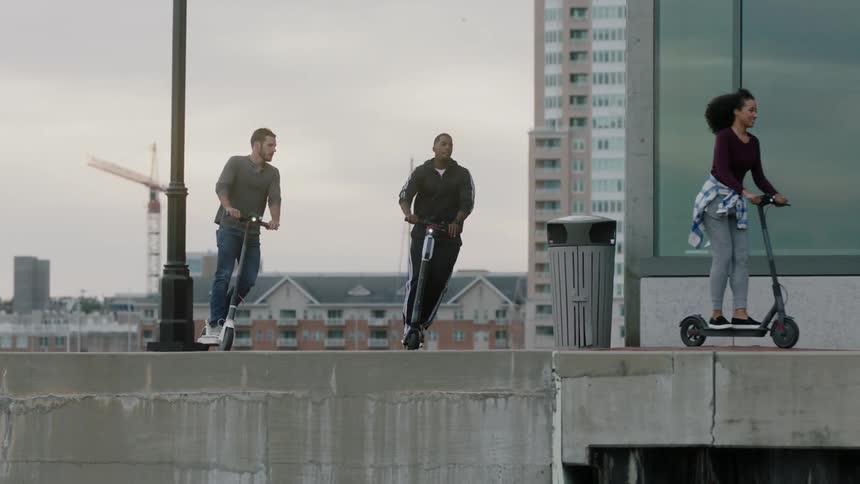 Friends ride scooters along a city building