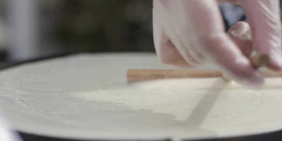 A chef uses a wooden tool to spread the batter evenly on the stone