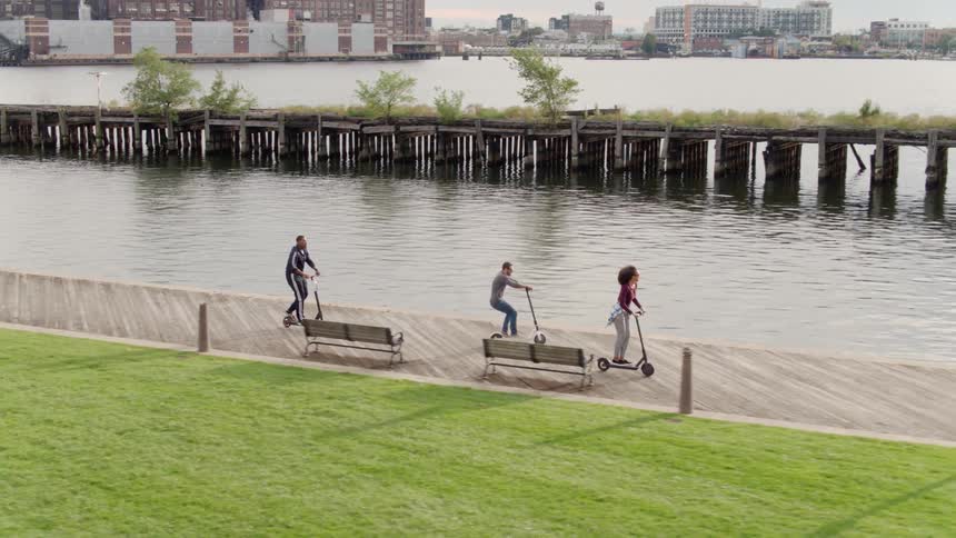 People riding kick scooters in an area next to some water in a city