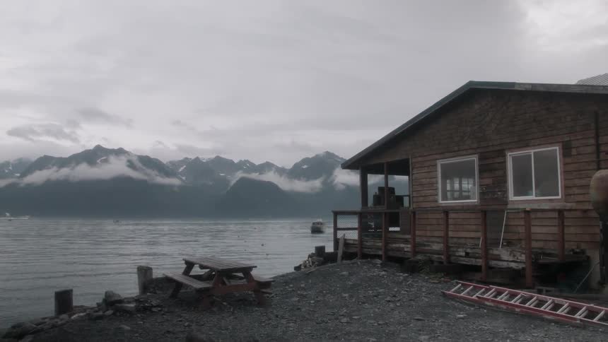 A boat is approaching a cabin on a shore in a mountain landscape on a cloudy day