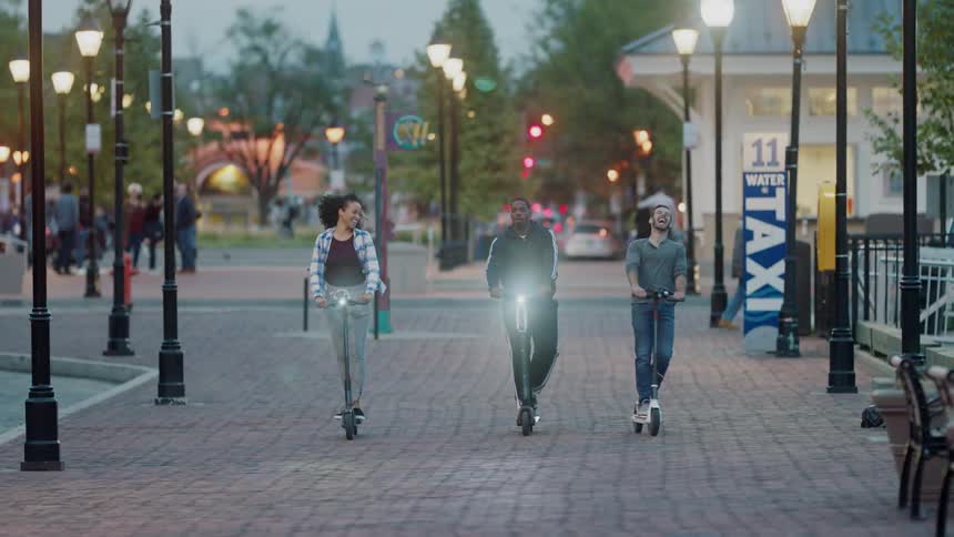 Three people are riding scooters down a busy city sidewalk
