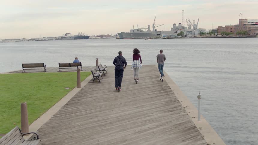 Friends are riding scooters to the end of a boardwalk in a city at dusk
