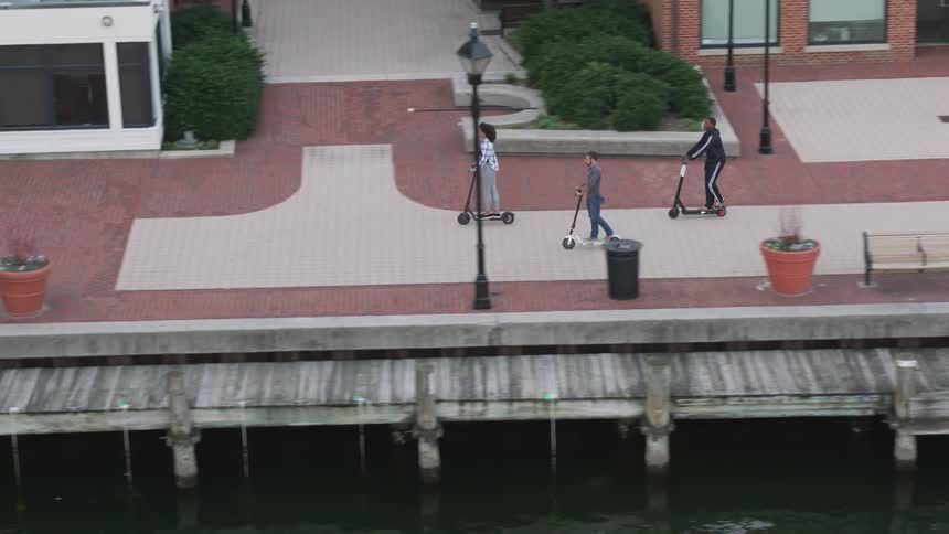 People riding kick scooters in an area next to some water in a city