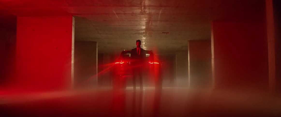A man stands in front of the red taillights of a car while in a parking garage