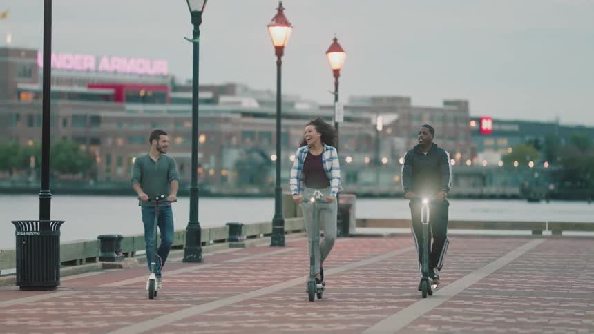 Friends are riding scooters along a boardwalk in a city at dusk
