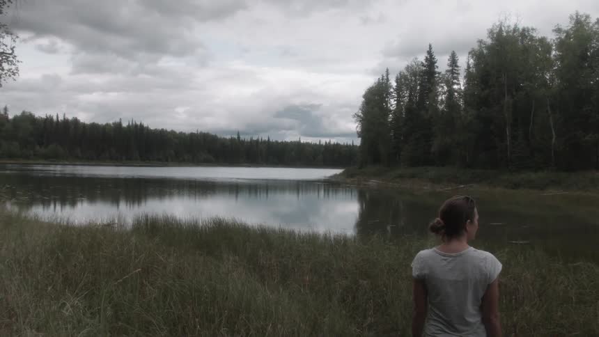 A woman stands out and looks towards a forest lake