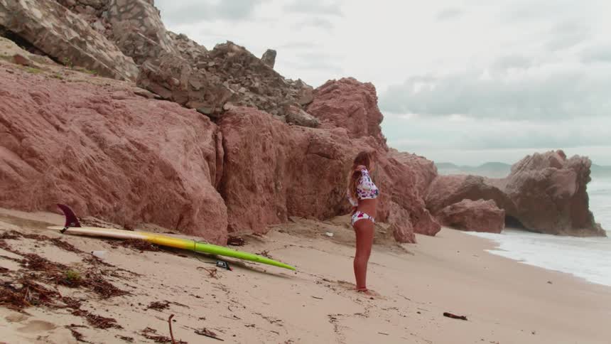 A woman is standing on the beach stretching with her surfboard behind her