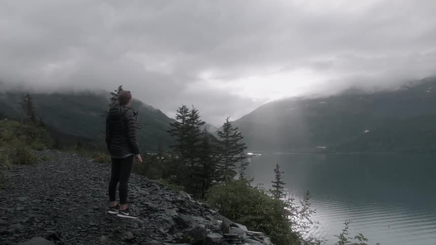 A woman stands on a rocky trail looking down at the water