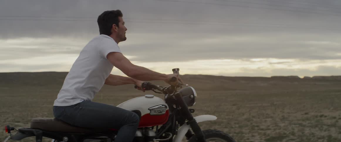 A man rides a motorcycle across a rural field at sunset