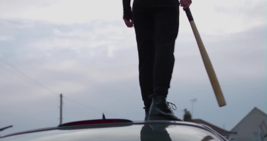A woman stands on a car roof with a baseball bat