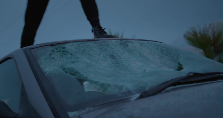 A person standing on a car breaks the windshield with a bat