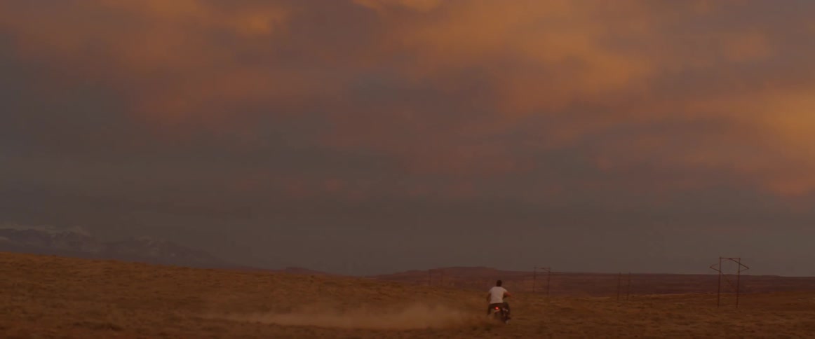 A man rides a motorcycle across a rural field at sunset