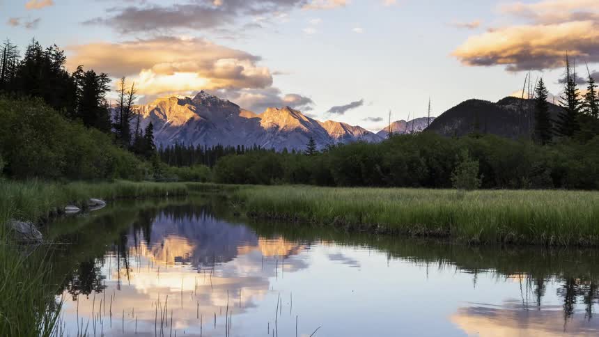 The calm lake water is reflecting the mountain and moving clouds