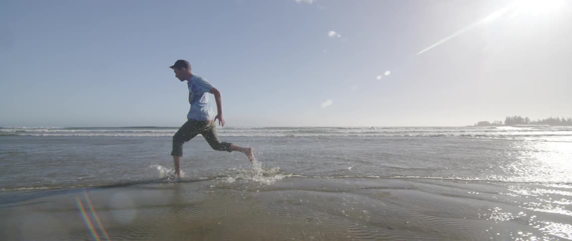 A young white man runs and hops along a beach