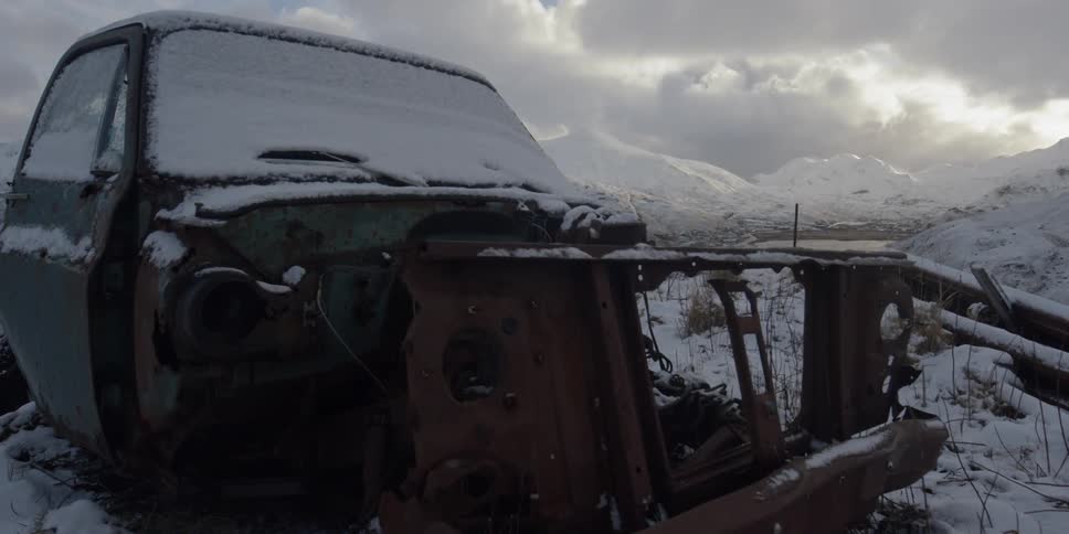 A rusted truck frame sits in snow and grass on a cloudy day