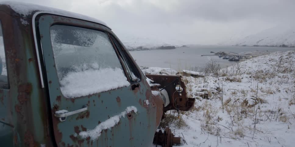 A rusted truck sits on the snowy ground by the water