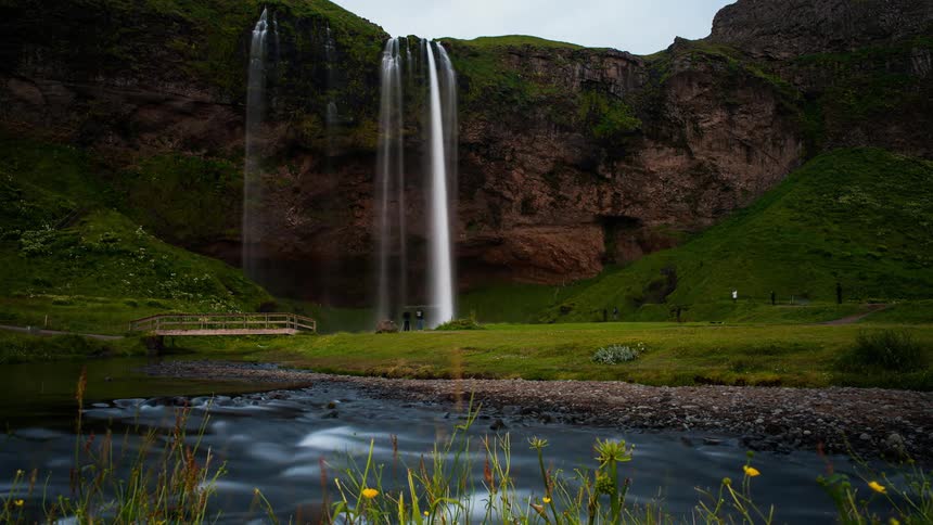 People are walking on a path near a waterfall as day passes to night