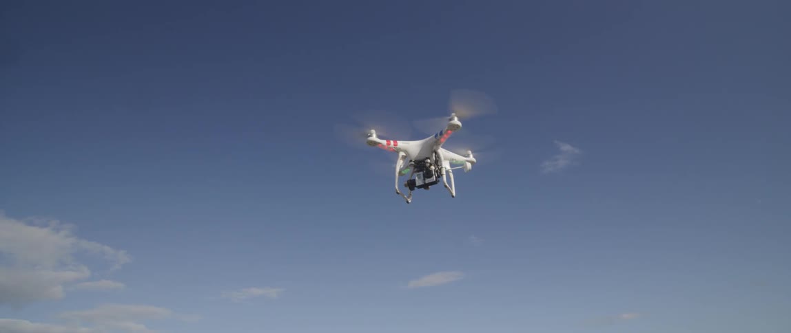 A drone flies in a partially cloudy sky